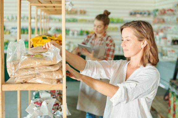 Femme range des paquets de produits sur une étagère dans un magasin. Une autre personne est en arrière-plan.