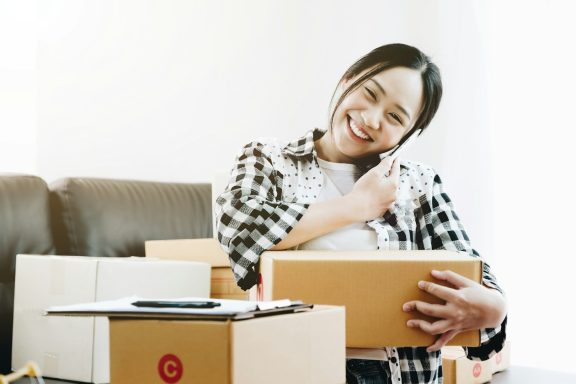 Femme souriante tenant une boîte, entourée de colis dans un intérieur lumineux.